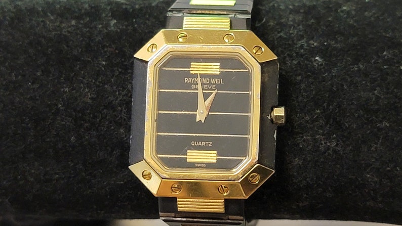 May include: A gold and black Raymond Weil Geneve quartz wristwatch with a black face and gold hands.
