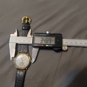 May include: A gold-tone wristwatch with a black leather band is being measured with a digital caliper. The caliper reads 34.89 mm.