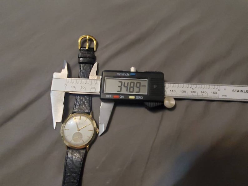 May include: A gold-tone wristwatch with a black leather band is being measured with a digital caliper. The caliper reads 34.89 mm.