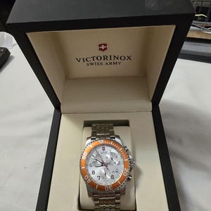 May include: A silver and orange chronograph wristwatch with a white face and a black box with the Victorinox Swiss Army logo.