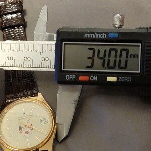 May include: A gold-toned wristwatch with a cream-colored face, a date display, and a brown leather band. The watch is measured at 34 mm using a digital caliper. The watch face features a logo and a small design.