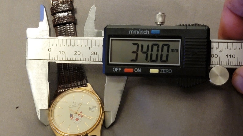 May include: A gold-toned wristwatch with a cream-colored face, a date display, and a brown leather band. The watch is measured at 34 mm using a digital caliper. The watch face features a logo and a small design.