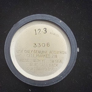 K&ouml;nnte beinhalten: Nahaufnahme der R&uuml;ckseite einer silbernen Uhr mit schwarzem Rand. Das Zifferblatt der Uhr tr&auml;gt den Text "12-3 3308 USE ONLY GENUINE ACCUTRON CELL, MARKED 218 INSTALL WITH YELLOW SEAL TOWARD CASE BACK (218 TOWARD DIAL)"
