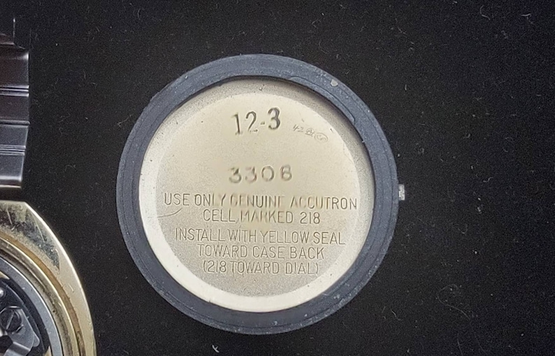 K&ouml;nnte beinhalten: Nahaufnahme der R&uuml;ckseite einer silbernen Uhr mit schwarzem Rand. Das Zifferblatt der Uhr tr&auml;gt den Text "12-3 3308 USE ONLY GENUINE ACCUTRON CELL, MARKED 218 INSTALL WITH YELLOW SEAL TOWARD CASE BACK (218 TOWARD DIAL)"