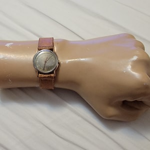May include: A gold-tone wristwatch with a white face and black hands. The watch has a brown leather band.