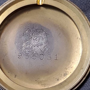 May include: Close-up of the back of a gold-toned watch. The watch has a circular case with a smooth, polished surface. The inscription "USSENARD" is visible in the center of the case, along with the number "955031".