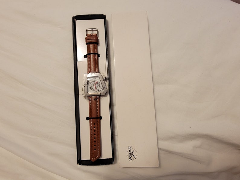 May include: A silver watch with a brown leather band in a black box. The watch face is a unique geometric shape with the words "Flight" and "World" printed on it. The box has the word "Sweda" printed on it.
