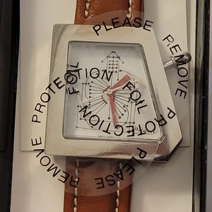 May include: A silver watch with a white face and a brown leather band. The watch face has a unique design with a red hand and the text "PLEASE REMOVE PROTECTION FOIL" printed on the clear plastic covering.