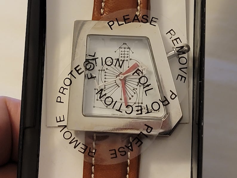 May include: A silver watch with a white face and a brown leather band. The watch face has a unique design with a red hand and the text "PLEASE REMOVE PROTECTION FOIL" printed on the clear plastic covering.