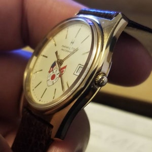 May include: Gold-toned Hamilton Masterpiece watch with a cream-colored face, featuring a date display and a logo with red and blue accents. The watch has a brown leather strap.