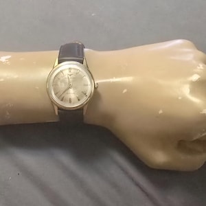 May include: A gold-tone analog wristwatch featuring a round face with a silver dial, gold-colored hour markers, and thin hands. The watch is equipped with a dark brown leather strap that has a smooth texture and a standard buckle closure mechanism.