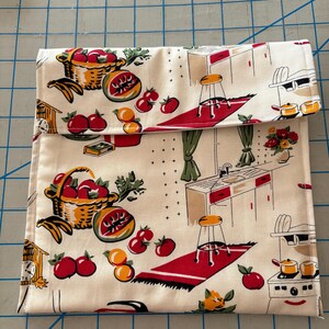 Puede incluir: Un envoltorio rectangular para sándwich de color crema con un estampado retro de cocina y frutas. La tela presenta ilustraciones de tomates rojos, naranjas y una cesta de manzanas. El envoltorio está doblado.