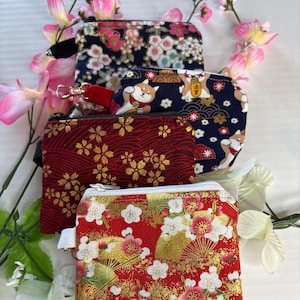 May include: Four small, zippered fabric pouches with Japanese-inspired patterns. The pouches feature floral and Shiba Inu dog designs in red, navy, and gold. The pouches are arranged with pink and white flowers.