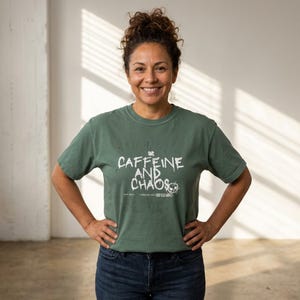May include: A green t-shirt with the words "CAFFEINE AND CHAOS" printed in white. The shirt has a distressed look and is paired with blue jeans. The person is smiling and has their hands on their hips.