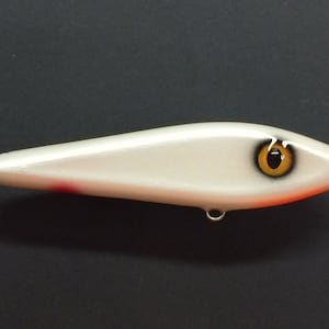 May include: White fishing lure with a black and orange eye. The lure has a red stripe on the bottom.