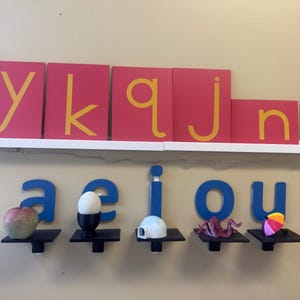 May include: Educational display featuring red letter cards with yellow letters and blue vowel letters. Small shelves hold a variety of objects, including a red apple, an egg in a holder, a small house, a purple octopus, and a colorful umbrella.
