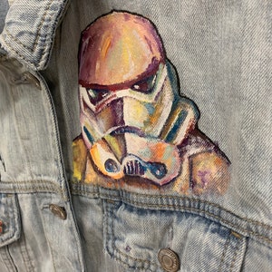 May include: A light blue denim jacket with a hand-painted image of a Star Wars Stormtrooper on the front. The Stormtrooper is painted in a colorful abstract style.