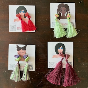 May include: Four pairs of handmade tassel earrings. Each pair features a unique design with heart-shaped accents and colorful tassels. The earrings are displayed on small cards with illustrations and the text "RUBY" and "SYNNA accessory".