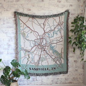 May include: A woven tapestry wall hanging featuring a map of Nashville, Tennessee. The map is in shades of brown, green, and blue, with the city name and state abbreviation printed in black at the bottom. The tapestry is hanging on a white brick wall.