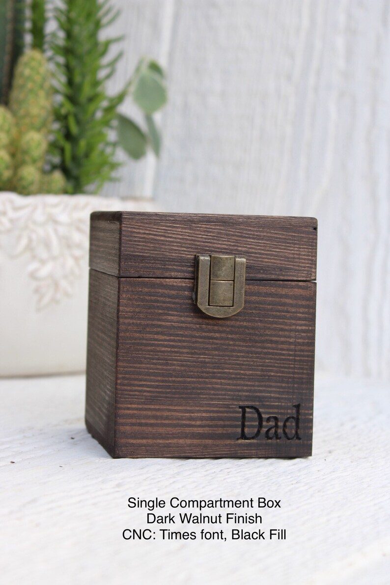 May include: A dark walnut wood box with a single compartment and a brass latch. The box is engraved with the word "Dad" in a Times font with black fill.