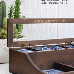 May include: A dark walnut valet box with a navy blue nautical map fabric lining. The box has a hinged lid with a clear glass insert and a brass hinge. The lid is engraved with the coordinates "N 35&deg; 41' 26.52" W 83&deg; 51' 27".
