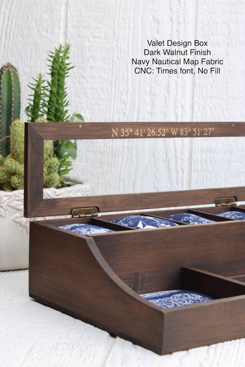 May include: A dark walnut valet box with a navy blue nautical map fabric lining. The box has a hinged lid with a clear glass insert and a brass hinge. The lid is engraved with the coordinates "N 35&deg; 41' 26.52" W 83&deg; 51' 27".
