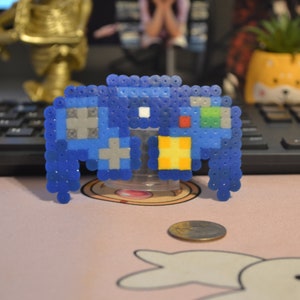 May include: A blue pixel art games controller made from small beads. The controller has grey, yellow, and red details. It is displayed on a clear stand, with a coin in the foreground. The background includes a keyboard and a small potted plant.