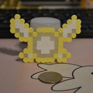 May include: A pixelated yellow and white charm with a cross design, resembling a video game character. A coin is next to the charm. The charm is made of small, round beads.