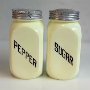 May include: Two pale yellow glass shakers with silver-toned metal lids. The left shaker features the word PEPPER printed in black, while the right shaker features the word SUGAR printed in black. Both containers have a rectangular shape.