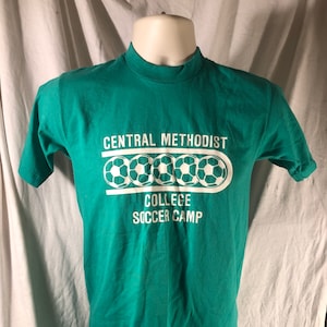 May include: A green short-sleeve t-shirt with a mock neck collar. The front features white text reading CENTRAL METHODIST COLLEGE SOCCER CAMP and a graphic of five footballs arranged in a horizontal row within a white outlined border.