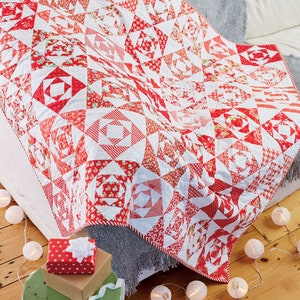 Könnte beinhalten: Eine rot-weiße Patchwork-Decke mit geometrischem Muster, drapiert über ein weißes Sofa mit einer grauen Decke. Die Decke ist von weißen Lichterketten und einem kleinen grünen Hocker mit einer roten Geschenkbox umgeben.