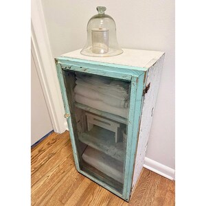 May include: A vintage cabinet with a glass door and shelves, painted white with turquoise trim. A glass cloche with a small jar sits on top. The cabinet is filled with folded white linens.
