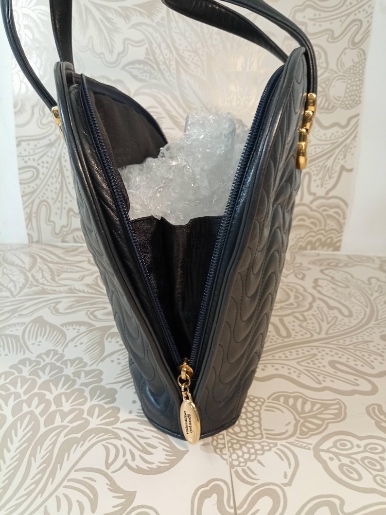 May include: A dark navy blue leather handbag featuring a quilted wave pattern on the exterior. The bag has a top zipper closure with a gold-toned metal pull tab engraved with the text Valentino Orlandi and two thin matching leather shoulder straps.