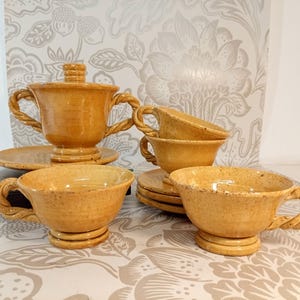 Op de afbeelding: Een keramische theeset in een warme, gespikkelde geelbruine tint. De set bevat kopjes, schotels en een suikerpot met deksel. De handvatten van de kopjes en de suikerpot zijn ingewikkeld ontworpen met een touwachtige textuur. De set staat op een oppervlak met bloemmotief.