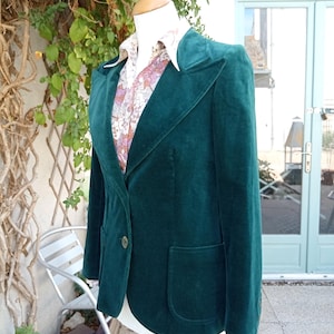 May include: A dark green velvet blazer featuring wide lapels, a single front button closure, and two patch pockets. The blazer is layered over a collared shirt with a light-coloured floral pattern in shades of pink, brown, and white.