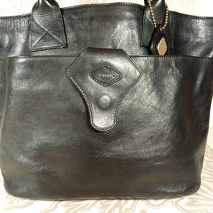 May include: A black leather tote bag with a flap closure and a silver metal handle. The bag has a diamond-shaped logo on the front that says "Tramontina".