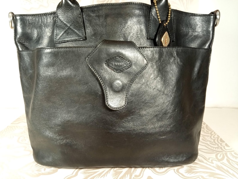 May include: A black leather tote bag with a flap closure and a silver metal handle. The bag has a diamond-shaped logo on the front that says "Tramontina".