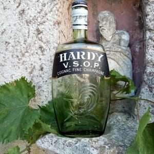 Könnte beinhalten: Eine grüne Glasflasche Hardy V.S.O.P. Cognac Fine Champagne. Die Flasche hat ein schwarzes Etikett mit weißem Text und einem Hahn-Emblem. Die Flasche ist teilweise gefüllt und hat ein weißes und schwarzes Etikett am Hals.