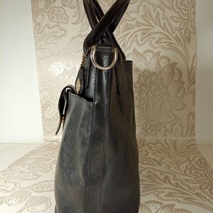 May include: A black leather tote bag with two top handles and a buckle closure. The bag has a front pocket and a silver-toned metal ring on the top handle.