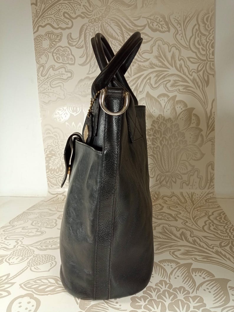 May include: A black leather tote bag with two top handles and a buckle closure. The bag has a front pocket and a silver-toned metal ring on the top handle.