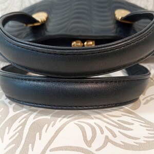May include: A black handbag featuring two rounded top handles and gold-toned metal hardware attachments. The surface of the bag displays a repeating embossed wavy pattern, and a gold-toned kiss-lock clasp is visible at the top center opening.