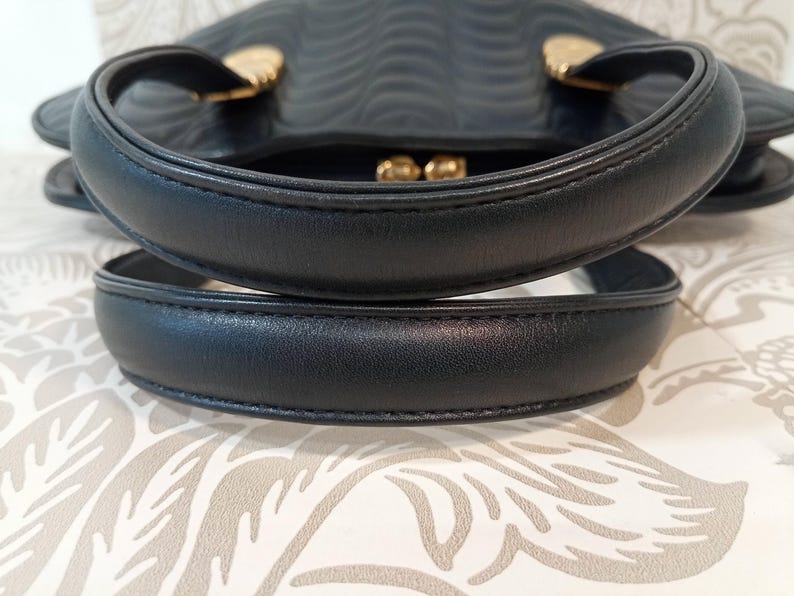 May include: A black handbag featuring two rounded top handles and gold-toned metal hardware attachments. The surface of the bag displays a repeating embossed wavy pattern, and a gold-toned kiss-lock clasp is visible at the top center opening.