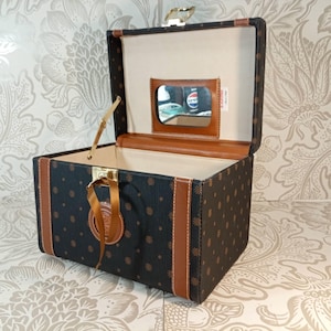 May include: A black vanity case with a brown repeating pattern, featuring tan leather-like trim, a gold-tone metal latch, and a rectangular mirror on the inside of the lid. The interior is lined with a light beige fabric and includes a small ribbon.