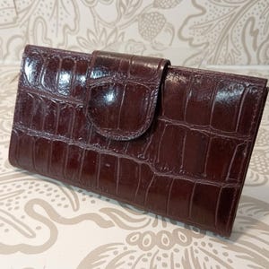 May include: A dark brown rectangular wallet with a glossy finish and a textured crocodile skin pattern. The wallet features a horizontal orientation and a central flap closure that secures the front panel with a rounded tab design.