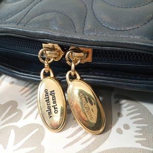 May include: A dark navy blue leather handbag featuring a quilted pattern and a black zipper closure. Two gold-tone oval metal zipper pulls are attached, with one engraved with the text Valentino Orlandi and the other with Made in Italy.