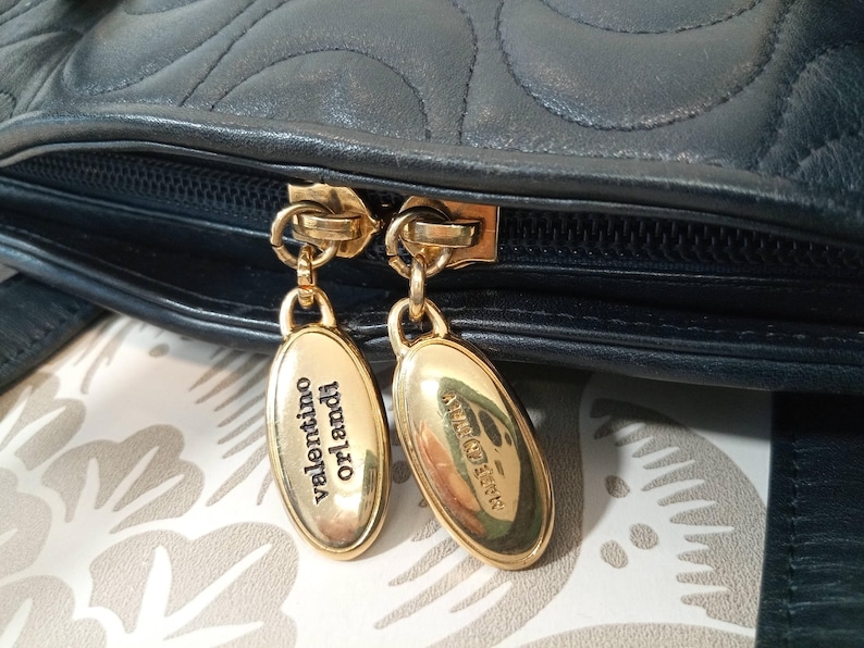 May include: A dark navy blue leather handbag featuring a quilted pattern and a black zipper closure. Two gold-tone oval metal zipper pulls are attached, with one engraved with the text Valentino Orlandi and the other with Made in Italy.