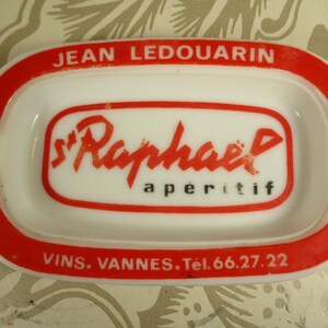 Peut inclure: Un cendrier en céramique blanche avec un bord rouge et des lettres rouges. Le cendrier porte le texte "Jean Ledouarin" en haut, "St Raphael apéritif" au centre et "Vins. Vannes. Tel. 66.27.22" en bas.