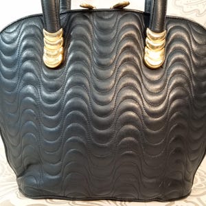 May include: A black handbag featuring a quilted wave pattern across the surface. The bag includes two top handles attached with gold-toned metal hardware. The top of the bag is secured with a zipper closure that features gold-toned metal pulls.