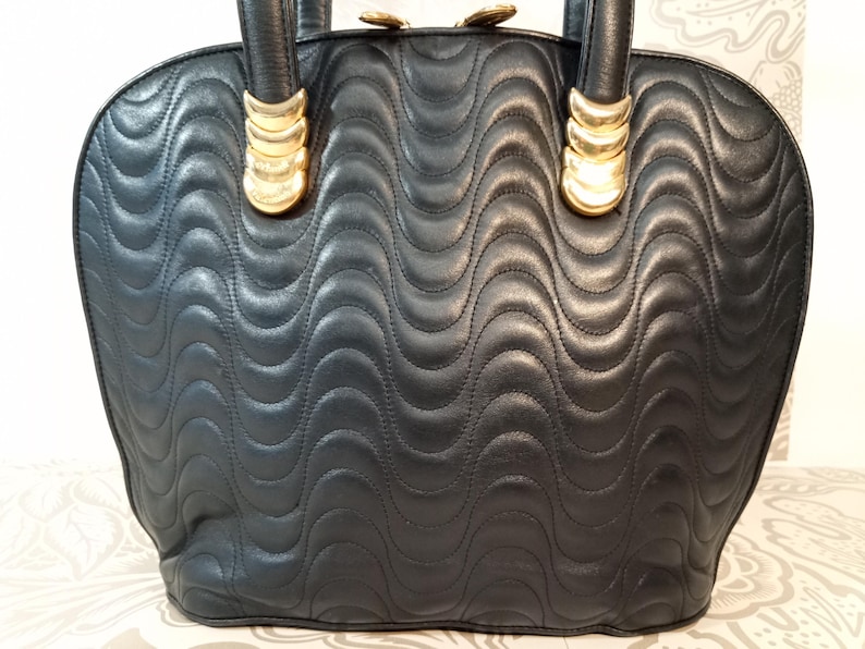 May include: A black handbag featuring a quilted wave pattern across the surface. The bag includes two top handles attached with gold-toned metal hardware. The top of the bag is secured with a zipper closure that features gold-toned metal pulls.