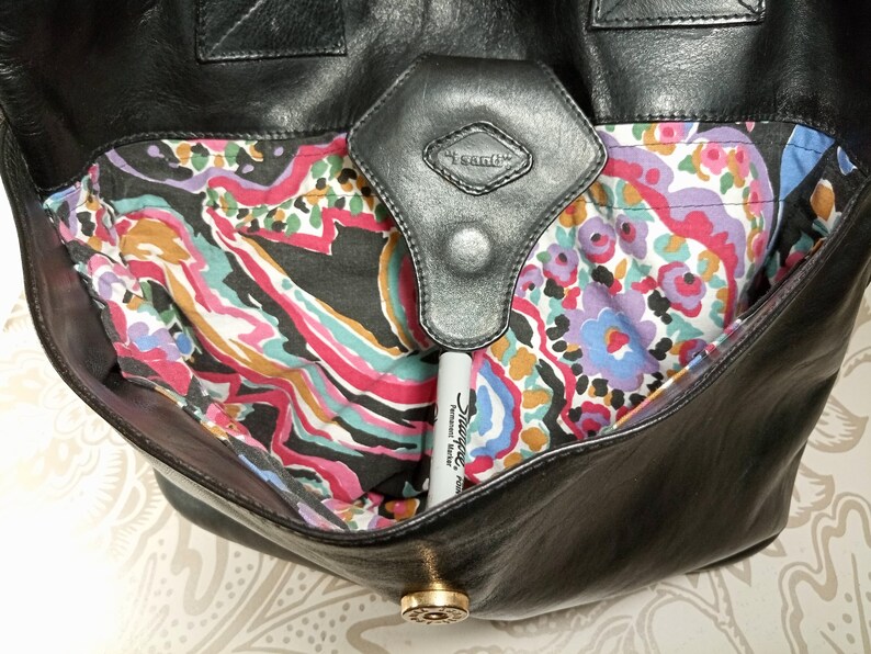 May include: Black leather tote bag with a paisley patterned lining. The bag has a flap closure with a metal button and a leather strap with a metal buckle.
